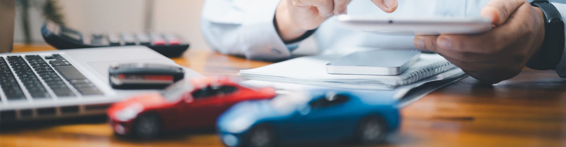 Man using a tablet at a wooden desk with a laptop, smartphone, notebooks, and blurred red and blue car models, suggesting work or study.