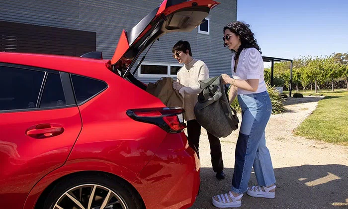 Red 2024 Subaru Impreza hatchback with rear boot open on a sunny day, two women loading backpacks into the cargo area